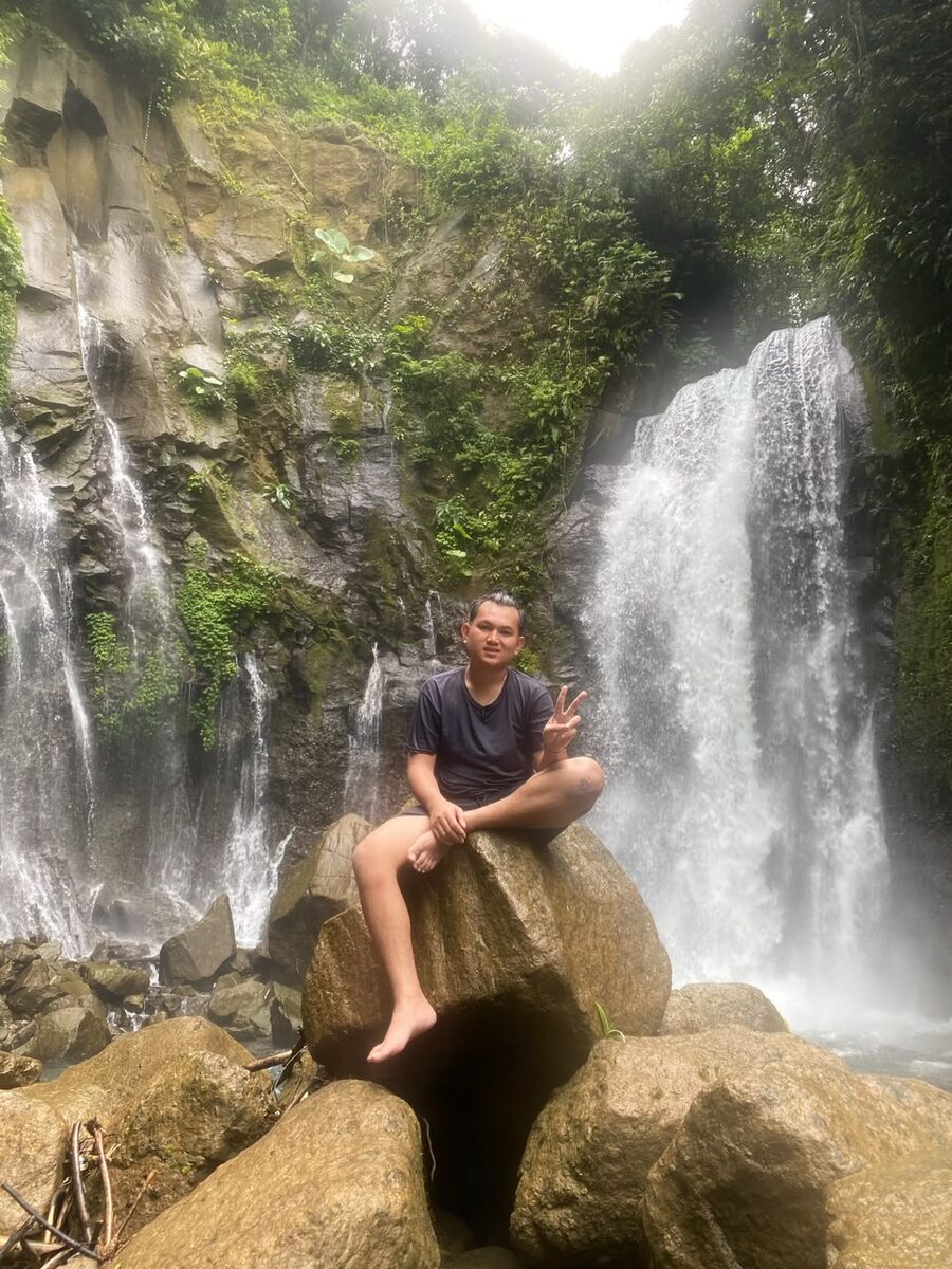 Duduk di atas batu besar dengan latar dua air terjun bertingkat