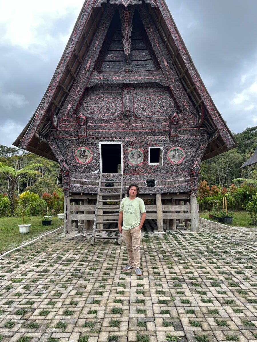 Berdiri di depan rumah adat Batak dengan atap ukiran khas