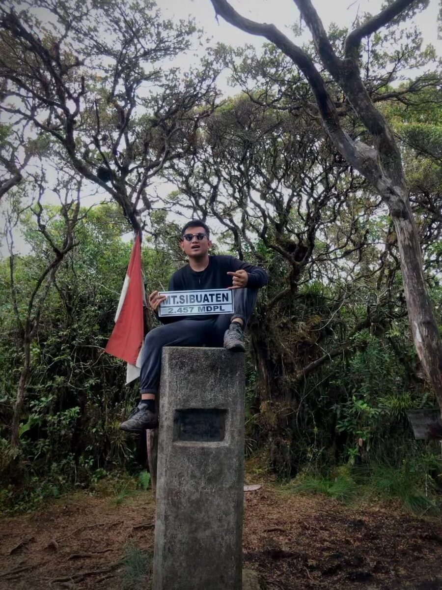 Duduk di atas tugu penanda puncak Gunung Sibuaten dengan bendera merah putih